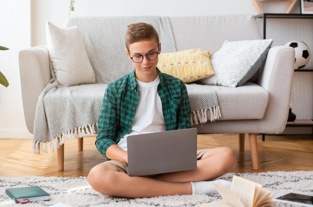 Smiling smart teenaged student working on school project at home, sitting on floor at his room, using laptop