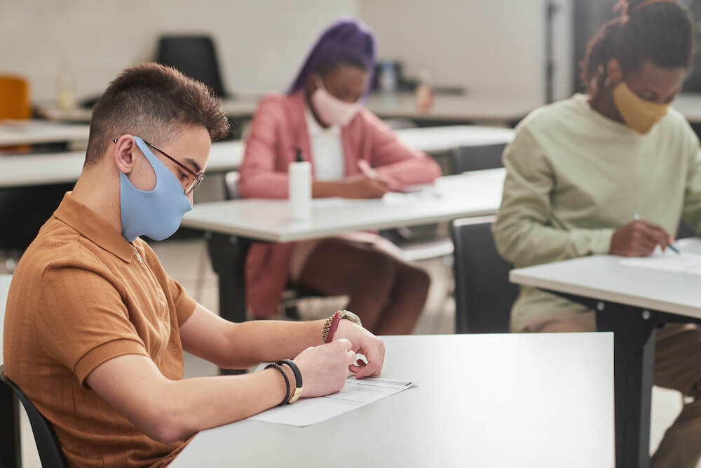 Side view portrait of young man wearing mask while taking test or exam in school with diverse group of people, copy space, Side view portrait of young man wearing mask while taking test or exam in school with diverse group of people, copy space