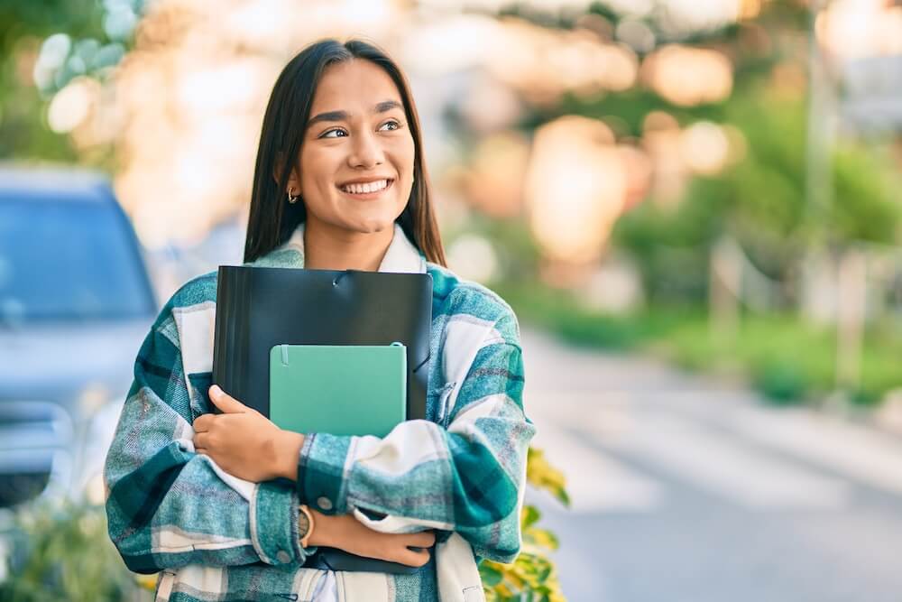 Young latin student girl smiling happy holding folder at the city.