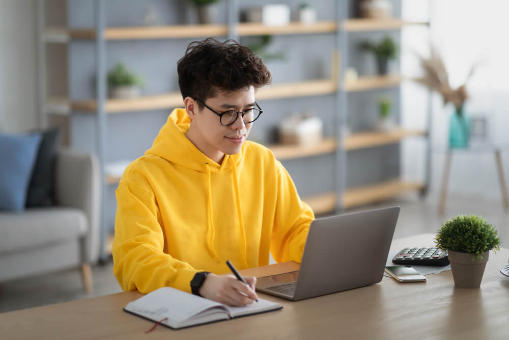 Distance Education Concept. Portrait of smiling Asian man in eyeglasses and yellow hoodie sitting at desk, using pc writing research, taking notes, watching tutorial or webinar studying online at home | Command Education