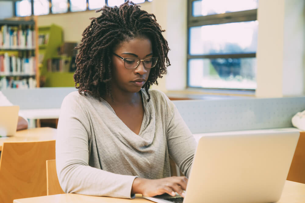 Focused student working in library computer class. Young black woman sitting at desk and using laptop in classroom with bookshelves