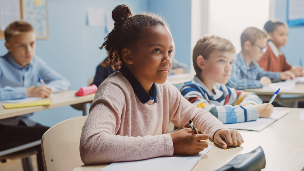 In Middle School Classroom Brilliant Black Girl Writes in Exercise Notebook, Taking Test and Writing Exam. Junior Classroom with Diverse Group of Children Working Diligently and Learning New Stuff