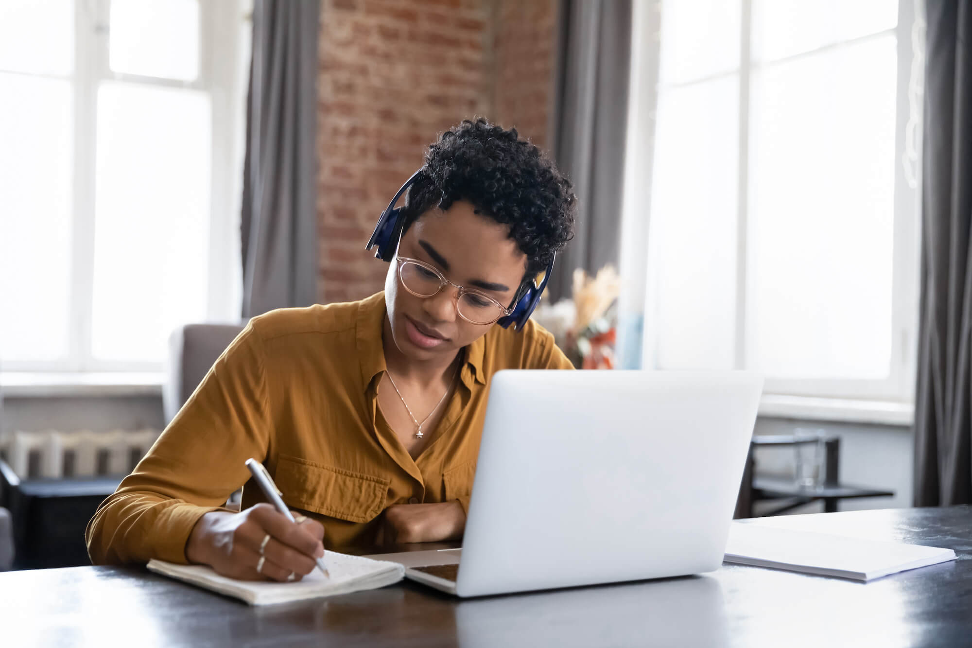 Focused student in glasses and headphones writing notes at laptop, checking handwriting summary, watching webinar, listening learning audio