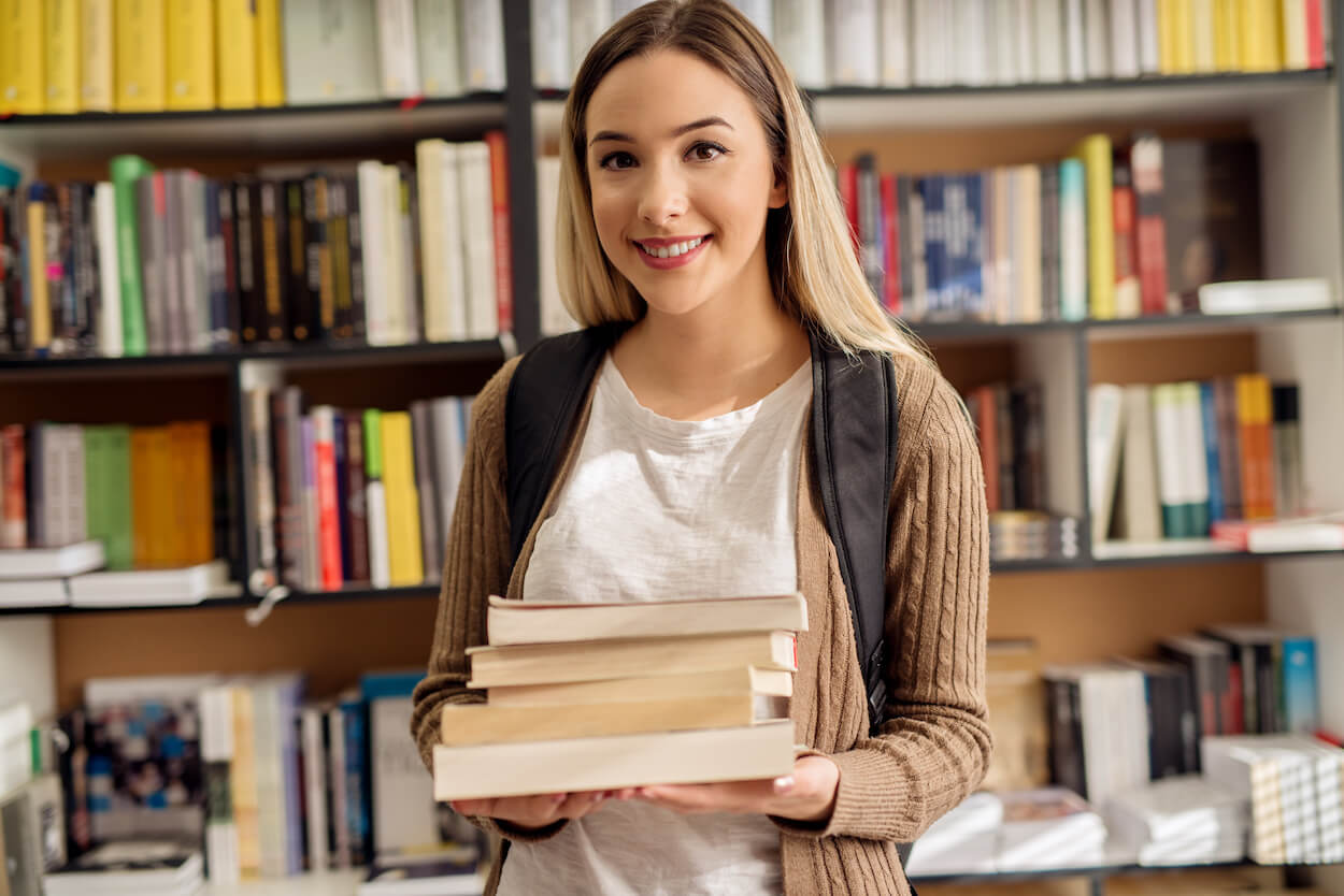 A smiling high school student girl standing with a backpack in the library while holding book stack in hands.