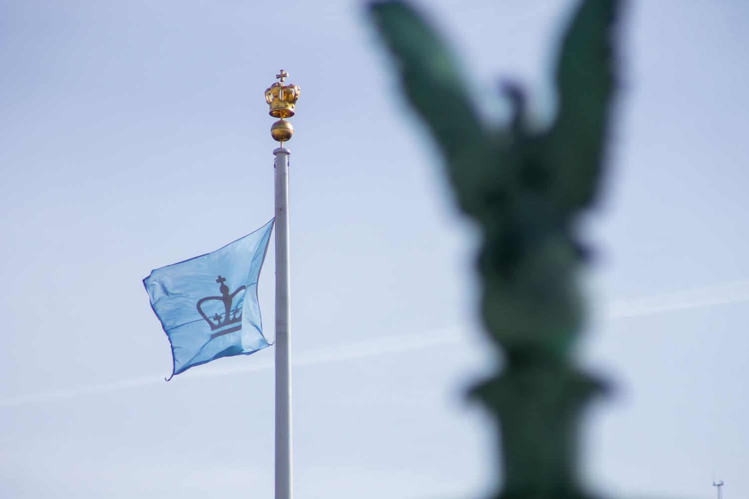 A light blue flag featuring a dark blue crown emblem waves in the wind atop a flagpole adorned with a golden crown finial. The background consists of a soft blue sky with wispy clouds, and the blurred silhouette of a statue is visible in the foreground.