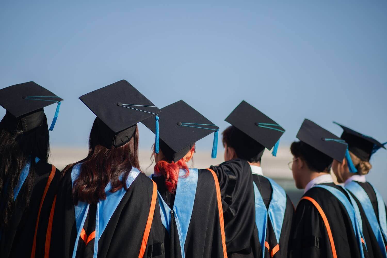A group of university graduates wearing black caps and gowns with blue tassels stand together, looking toward the horizon. The image represents academic achievement, higher education, and future aspirations.