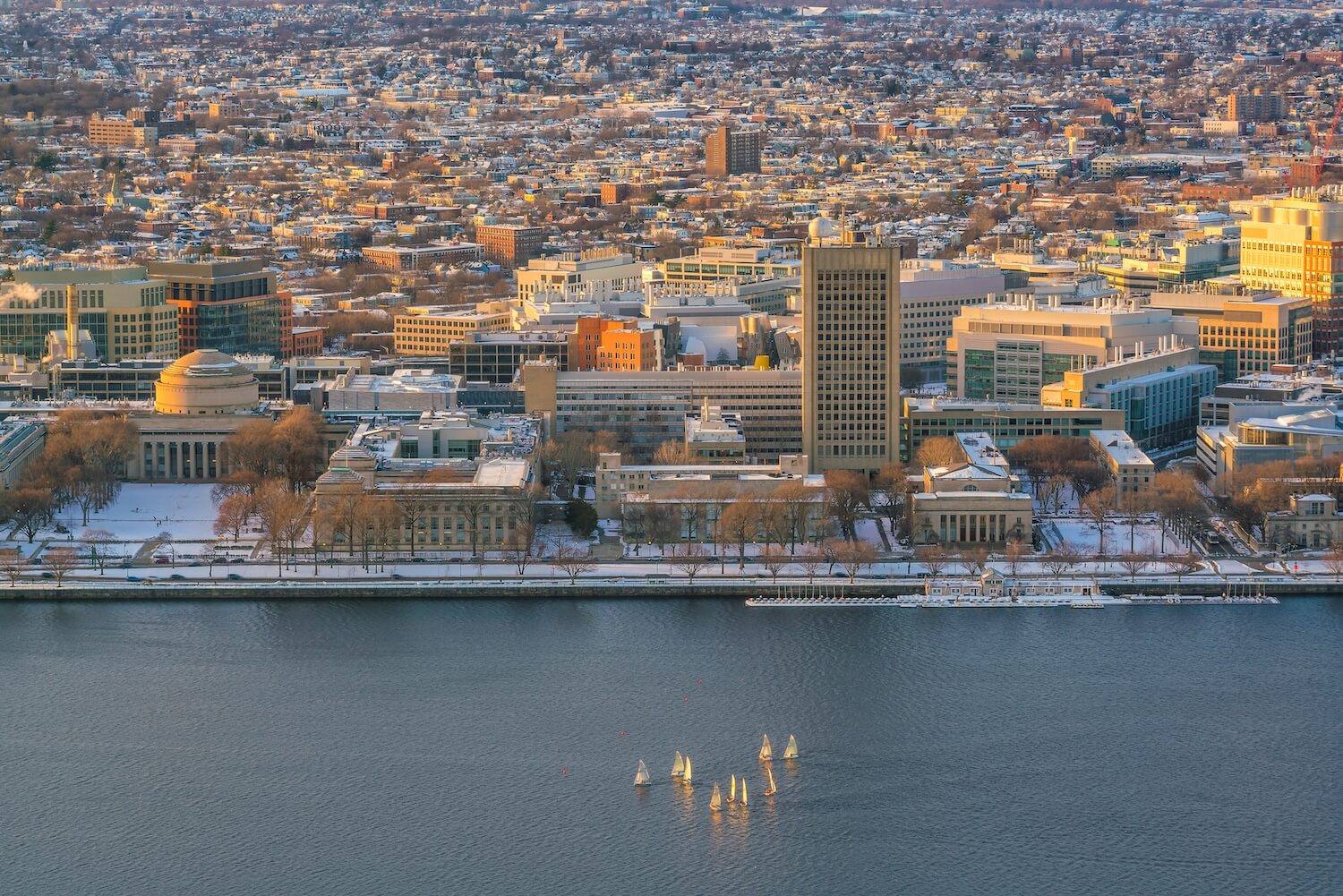 Aerial view of the Massachusetts Institute of Technology (MIT) campus in Cambridge, Massachusetts, with the Charles River in the foreground and the cityscape in the background. The image shows a winter scene with snow-covered buildings and small sailboats on the river.