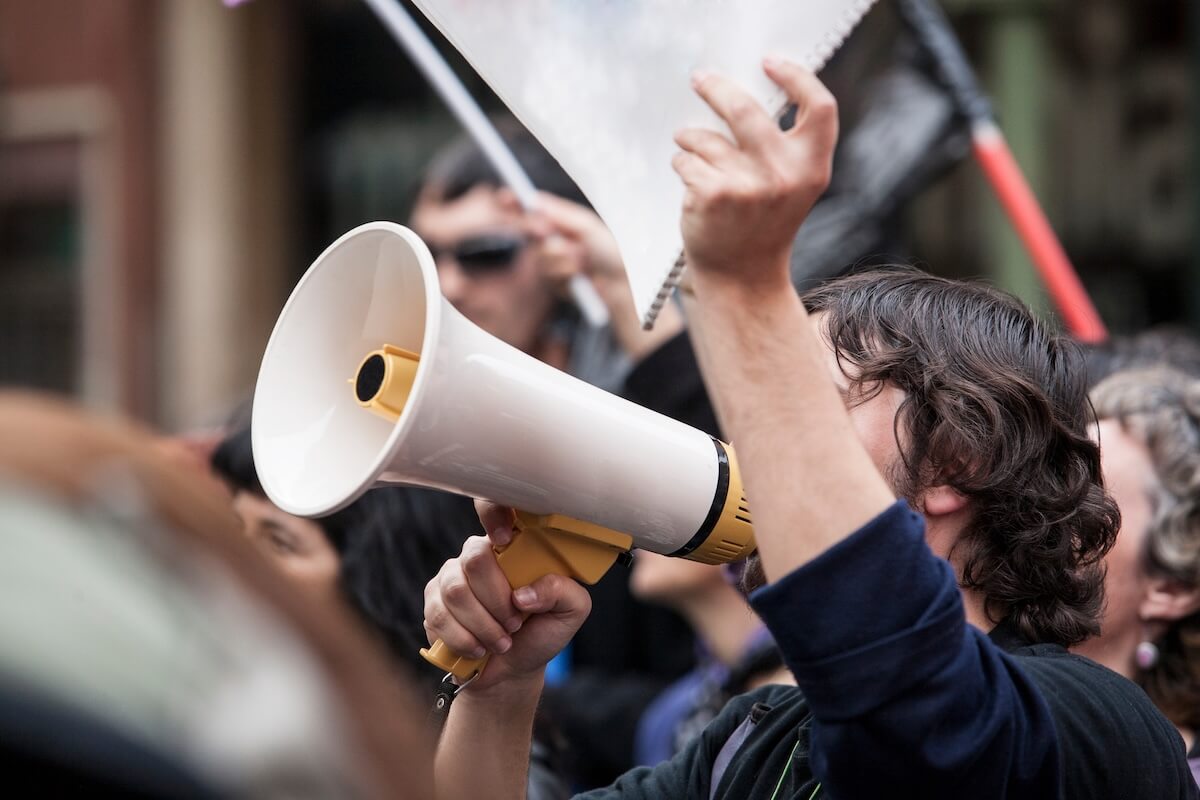 A protester holding a megaphone and raising a sign during a demonstration, surrounded by a crowd of people.