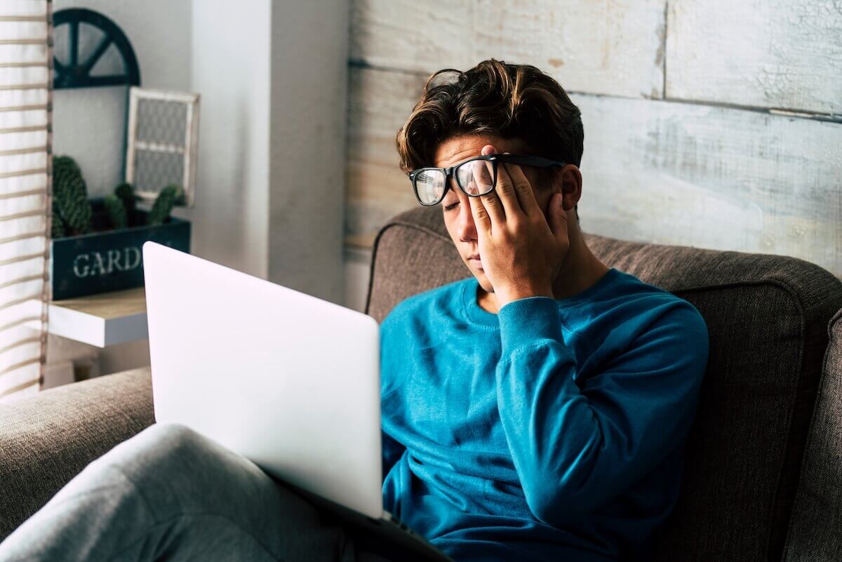 Stressed young entrepreneur sitting on a couch, rubbing his eyes in frustration while using a laptop, illustrating the pressure of college admissions despite academic and business success.