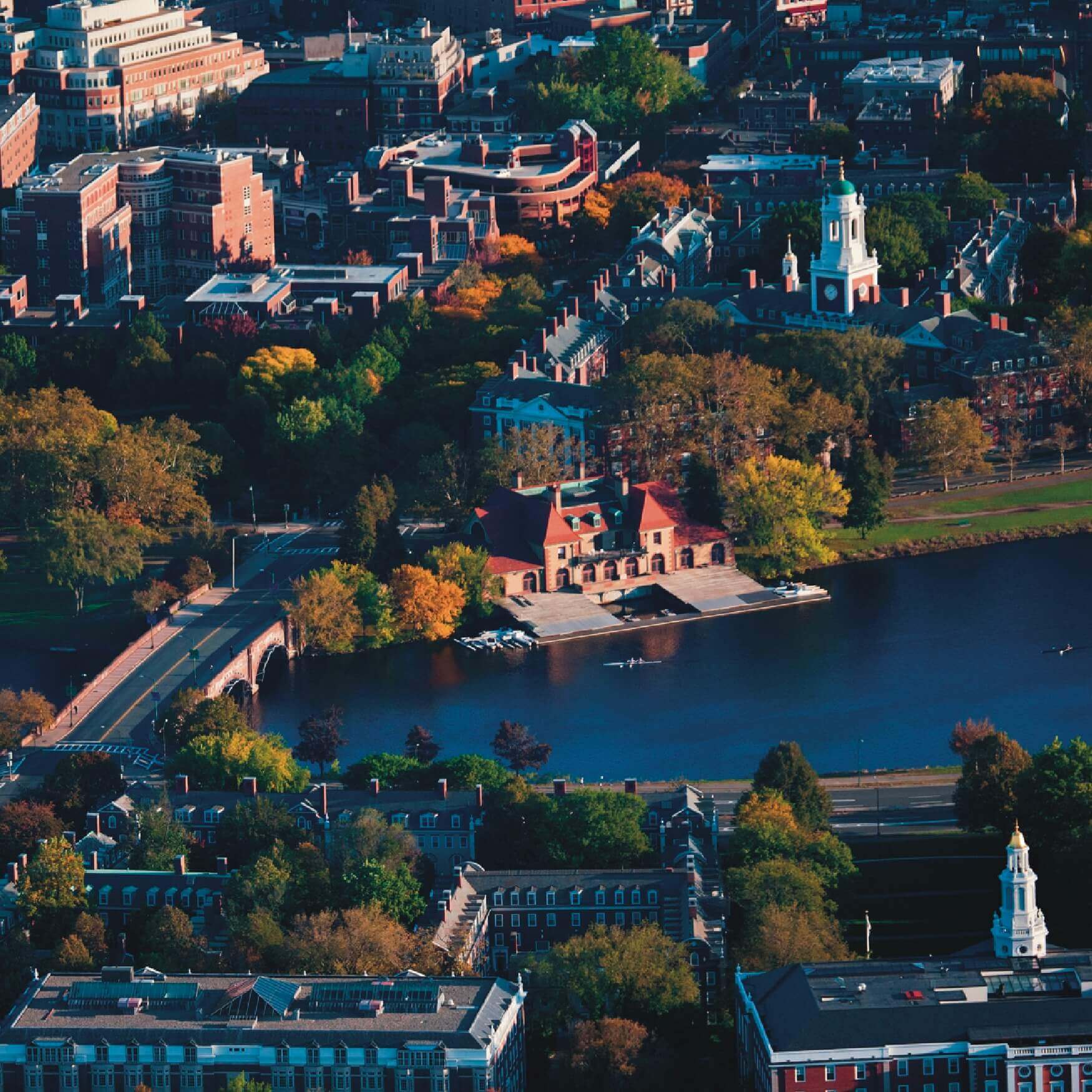 Aerial view of Harvard University’s Weld Boathouse, the Anderson Memorial Bridge, and the Charles River surrounded by fall-colored trees and historic Cambridge buildings.