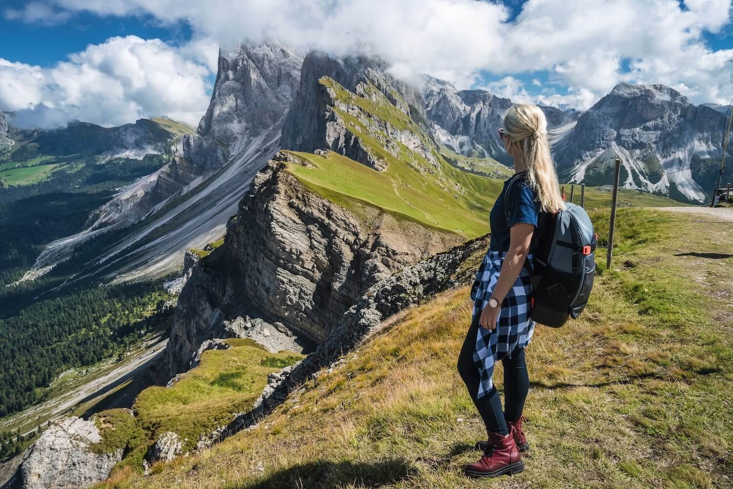 Young woman with a backpack stands at the edge of a cliff, overlooking the dramatic peaks of Seceda in the Dolomites during a gap year adventure in Italy.
