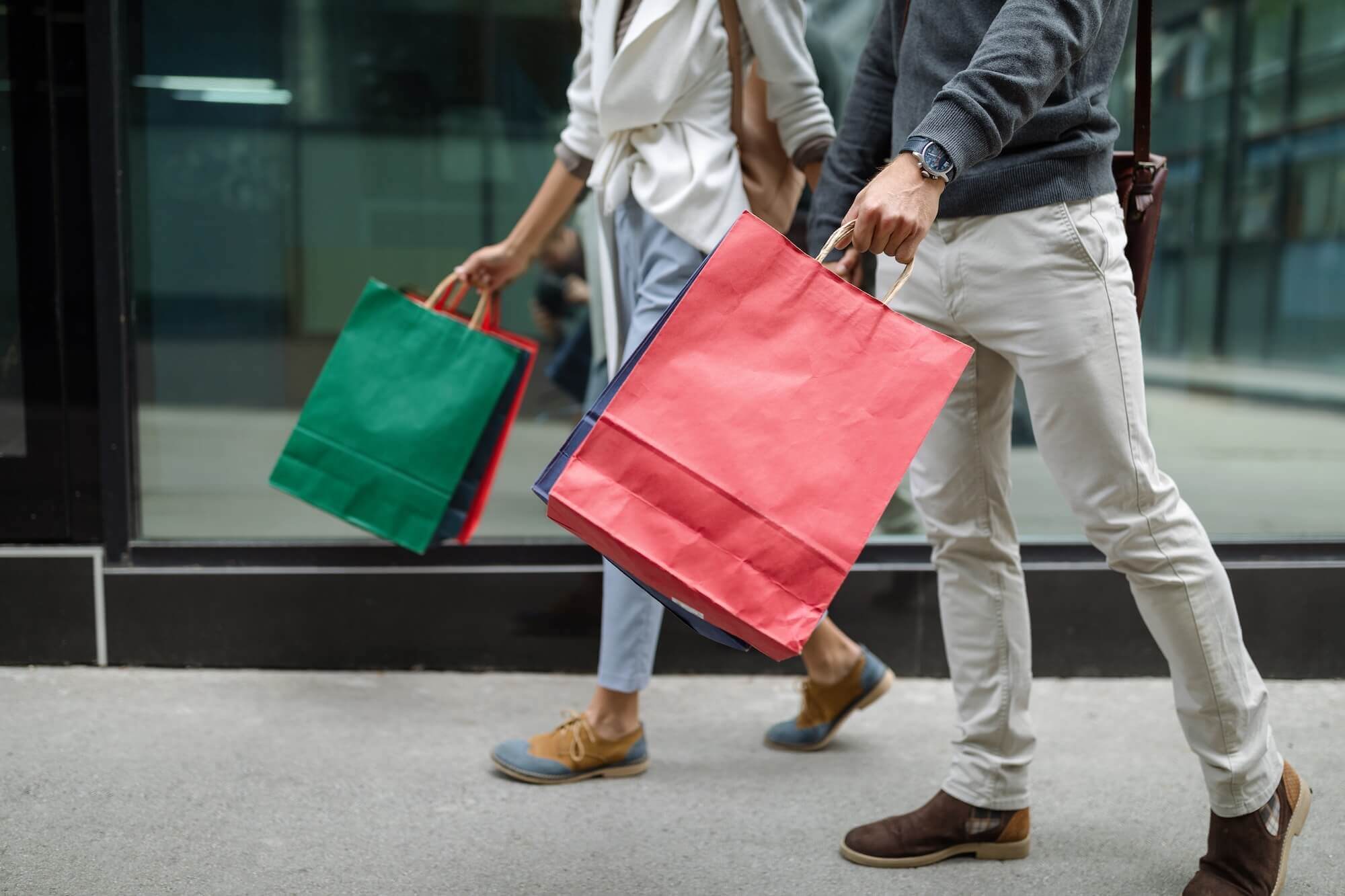 Two smiling shoppers walk down a city sidewalk carrying colorful shopping bags, reflecting renewed consumer confidence and economic optimism.