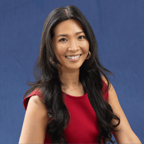 Grace Lee, Managing Director, wearing a sleeveless red dress, smiles warmly while sitting in a cozy, bright room