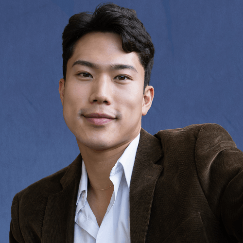 Min, Senior Mentor, wearing a brown blazer and white shirt, sitting on a bench with trees in the background