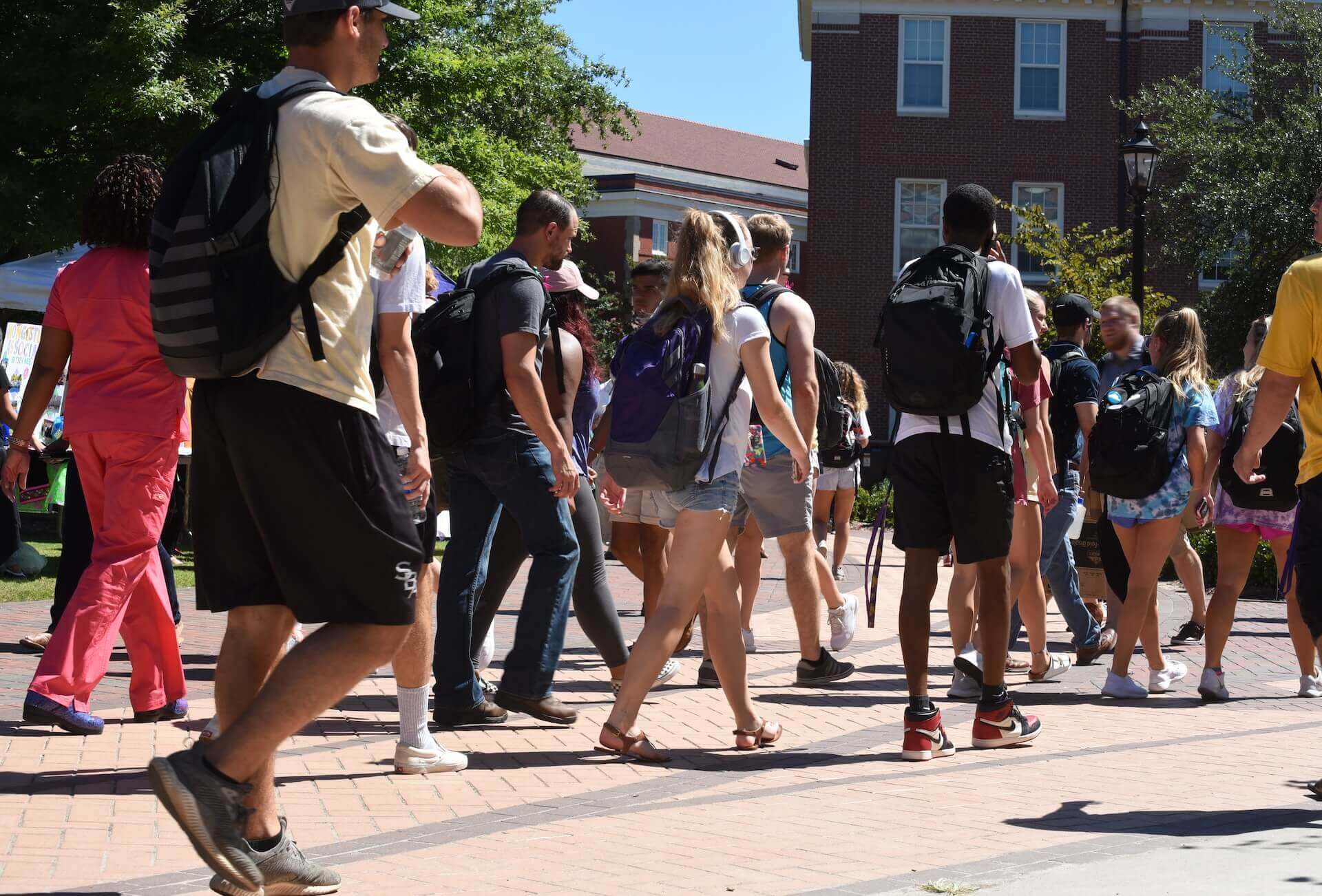 Diverse group of college students walking across brick campus courtyard with backpacks between red brick academic buildings on sunny day