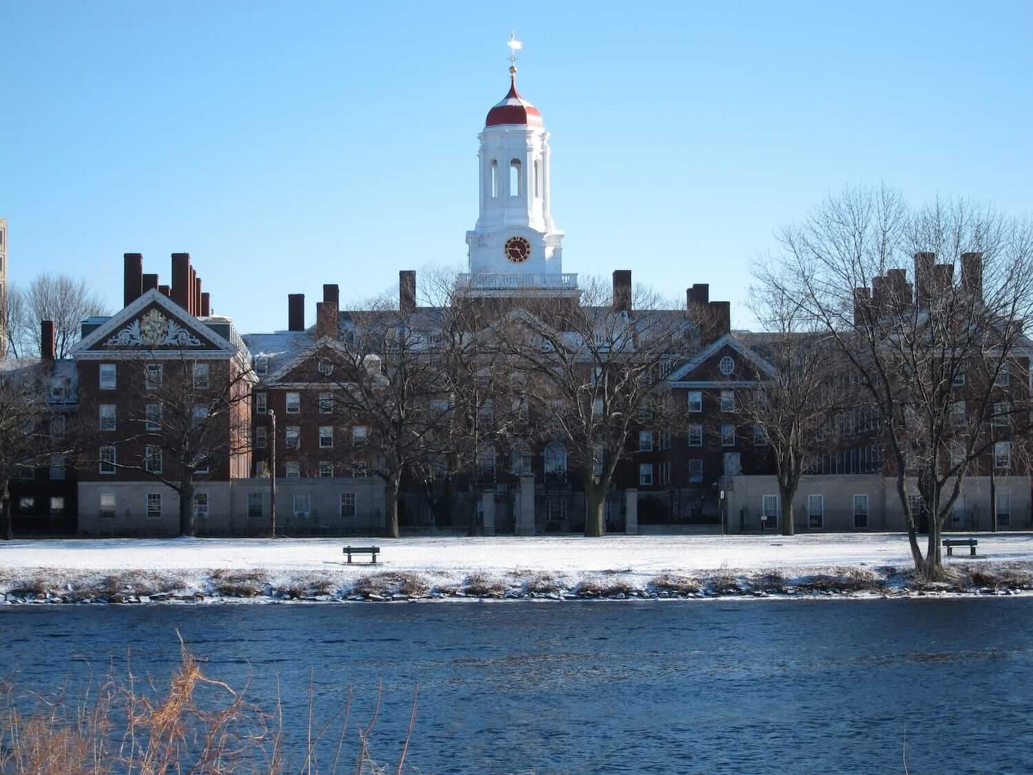 Harvard University campus viewed across the Charles River in winter, with snow-covered grounds and red-domed clock tower.