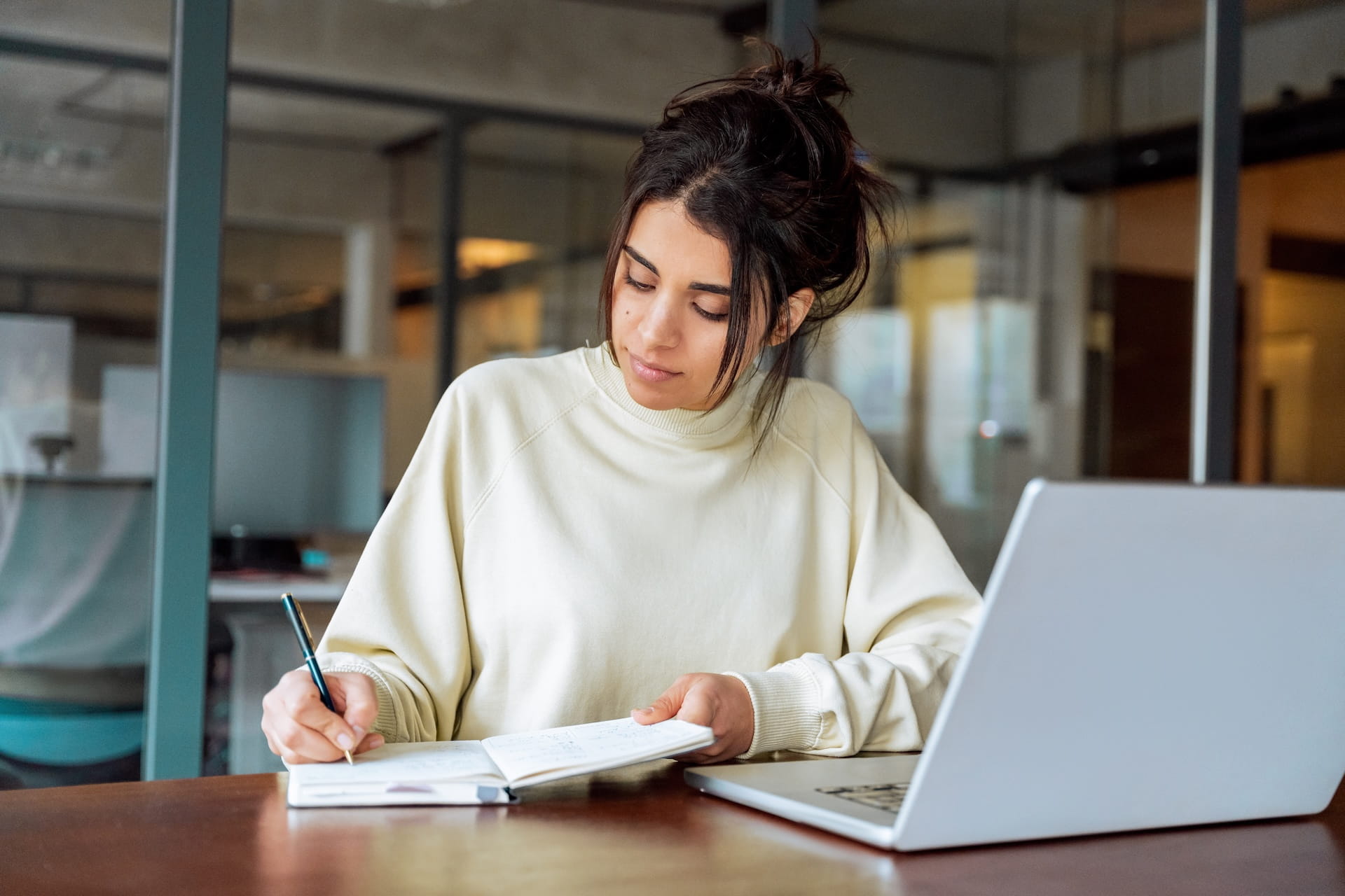 Happy graduate student with a certificate after graduation ceremony at the university. A college-aged woman with dark hair in a bun writes in a notebook at a desk with an open laptop in a modern workspace.