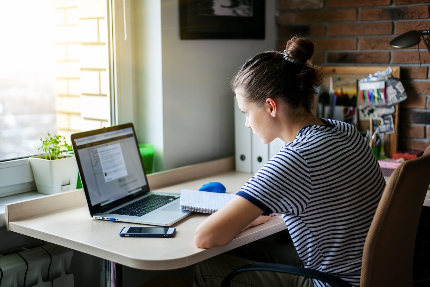 High school student in a striped shirt works at a laptop by a bright window, with a notebook and phone on the desk; calm, focused home office.